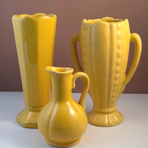 May include: Three yellow ceramic vases. The tallest has a scalloped rim and a hexagonal base. The middle vase has a small handle and a round body. The third vase has two handles and a decorative pattern. The vases are in a range of sizes.