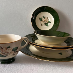 Puede incluir: Un juego de taza y platillo vintage de color crema con un diseño floral. El juego incluye una taza, un plato pequeño, un cuenco y dos platos más grandes. Los bordes están adornados con dorado y los platos tienen un borde verde oscuro.