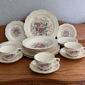 Peut inclure: Un service de table couleur crème orné d'un motif floral coloré composé de fleurs roses, jaunes, violettes et bleues avec des feuilles vertes. L'ensemble comprend plusieurs assiettes, bols et tasses avec soucoupes, finis par un liseré doré.