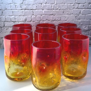 Pode incluir: Um conjunto de nove copos cilíndricos com um gradiente de cor vibrante que transita de um vermelho profundo no topo para um amarelo brilhante na base. Cada copo possui um acabamento de vidro texturizado e craquelado na superfície.