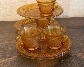 Könnte beinhalten: Ein Set aus bernsteinfarbenen Teetassen und passenden Untertassen aus Pressglas mit detaillierten floralen und geometrischen Prägemustern. Das Set besteht aus mehreren Tassen mit Henkeln und runden Tellern mit gewelltem Rand.
