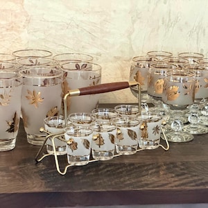 Könnte beinhalten: Ein Set aus mattiertem Glas mit Goldblattmotiven, bestehend aus hohen Trinkgläsern und Kelchgläsern. Ein Metallständer mit Holzgriff enthält acht kleine Schnapsgläser, die alle mit passenden Goldblattmustern verziert sind.