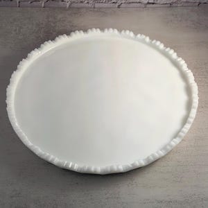 Puede incluir: Una fuente blanca de forma ovalada con un borde festoneado texturizado. La fuente está hecha de un material brillante y se encuentra sobre una superficie gris. El fondo presenta una pared de ladrillos blancos.