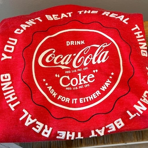 Op de afbeelding: Rood t-shirt met een wit Coca-Cola logo in een cirkel, en de tekst "YOU CAN'T BEAT THE REAL THING" rond de rand. Het shirt bevat ook de woorden "DRINK Coca-Cola" en "Coke" in het wit.