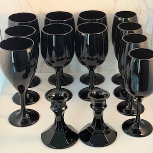 May include: A collection of ten black glass wine glasses with smooth, rounded bowls and slender stems, arranged in rows, accompanied by two black glass candle holders featuring faceted, geometric bases and wide, circular rims for candles.