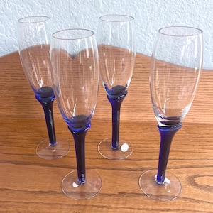 May include: Four clear champagne flutes with dark blue stems and bases. The stems have a unique, curved design. The glasses are arranged on a wooden surface, with a light-colored wall in the background.