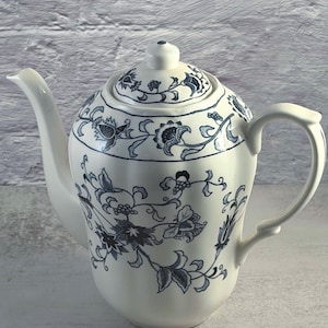 Puede incluir: Tetera de cerámica blanca con tapa, boquilla y asa. La tetera está decorada con un estampado floral azul. El diseño incluye flores, hojas y vides. La tetera está sobre una superficie gris claro.