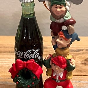 Op de afbeelding: Een Coca-Cola fles met een kerstkrans en een stapel van drie kabouterbeeldjes. De kabouters dragen rode, groene en blauwe outfits. Boven de kabouters bevindt zich een flesopener.