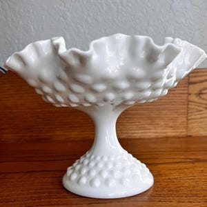 May include: A white hobnail glass pedestal bowl with a ruffled rim. The bowl has a round base and a stem. The bowl is made of white glass and has a textured surface. The bowl is likely vintage and suitable for serving desserts or displaying decorative items.