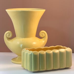 May include: Two yellow ceramic vases. One is a tall, urn-shaped vase with scrolled handles. The other is a rectangular planter with a scalloped rim and vertical ribbed sides. Both are on a white surface.