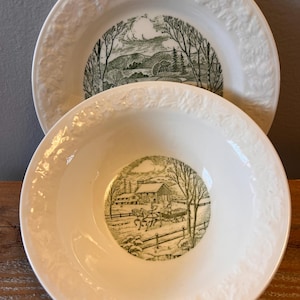 Może przedstawiać: Dwie białe ceramiczne miski z zielono-czarną sceną pasterską pośrodku. Miski mają dekoracyjny kwiatowy wzór na obrzeżach. Jedna to płytki talerz, druga głębsza miska.