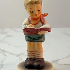 May include: A ceramic figurine of a young person with blonde hair, wearing a white shirt, orange neckerchief, and green shorts. The figure stands on a circular base, holding an open book with a red cover, looking down with closed eyes.