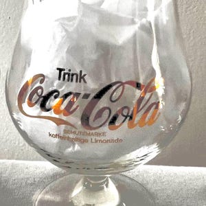 Op de afbeelding: Helder Coca-Cola glas met een gouden rand en steel. Het Coca-Cola logo is gedrukt in rood en zwart met het woord "Trink" erboven en "SCHUTZMARKE koffeinhaltige Limonade" eronder. Het glas is gevuld met wit vloeipapier.