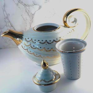 May include: A white porcelain teapot featuring gold-toned trim on the spout, handle, and lid, decorated with a delicate gold leaf garland pattern. The set includes a matching removable lid and a cylindrical white tea infuser with gold rim.