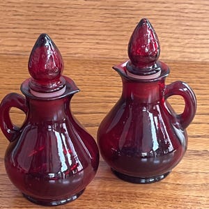 Puede incluir: Dos botellas de vidrio rojo rubí con tapones a juego. Cada botella tiene un asa pequeña y un pico. Los tapones tienen forma de lágrima. Las botellas están sobre una superficie de madera.