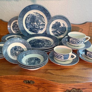 Op de afbeelding: Een verzameling blauw en wit keramisch servies, waaronder borden, kommen en theekopjes met schotels. Elk stuk heeft een gedetailleerde landschapsafbeelding. De set is op een houten oppervlak geplaatst.