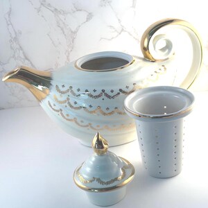 May include: A white porcelain teapot featuring a gold-trimmed spout, handle, and lid, decorated with a delicate gold leaf garland pattern. Beside the teapot sits a matching white porcelain tea infuser basket with a gold-rimmed top edge.