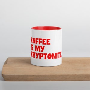 May include: White ceramic mug with a red rim and a red graphic that says "Koffee is my Kryptonite" with a coffee bean icon.
