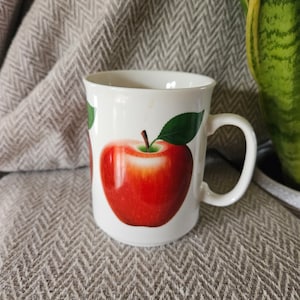 May include: White ceramic mug with a red apple design. The apple has a green leaf and stem. The mug has a handle.