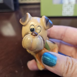 May include: A small, hand-painted ceramic figurine of a dog with brown and black fur. The dog has one eye closed and is holding a small, orange object in its mouth.