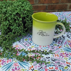 May include: A green and white ombre ceramic mug with the text "Java Momma" in black cursive writing. The mug is sitting on a white tablecloth with a colorful floral pattern.