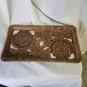 May include: A rectangular wooden tray featuring an intricate openwork carving of two large, rounded flower heads surrounded by detailed, swirling acanthus-style leaves. The border of the tray is defined by a repeating beaded or scalloped edge.