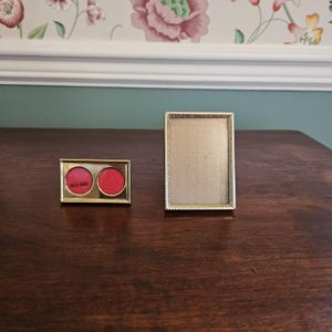 May include: Two vintage gold-toned metal frames on a dark wood surface. One is a rectangular picture frame, the other is a small compact with two red circles. The compact has the words "SOLID BRASS" printed on it.