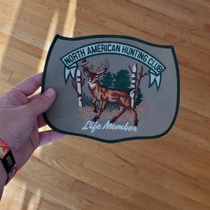 May include: A tan and green embroidered patch with a white deer in a forest scene. The patch reads "North American Hunting Club" and "Life Member".