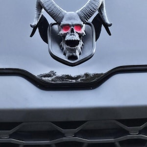 May include: A dark gray skull emblem with red eyes and large curved horns, mounted on a black vehicle. The skull has an open mouth and is set against a shield-shaped background.