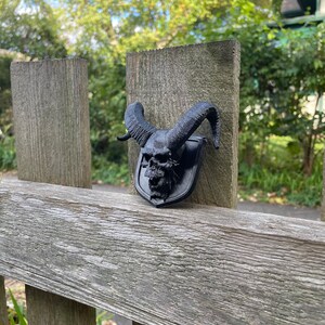 以下が含まれることがあります： 木製のフェンスの支柱に取り付けられた、角のある黒い3Dプリントの頭蓋骨。頭蓋骨は盾の形をしています。