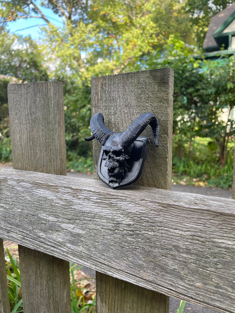 以下が含まれることがあります： 木製のフェンスの支柱に取り付けられた、角のある黒い3Dプリントの頭蓋骨。頭蓋骨は盾の形をしています。