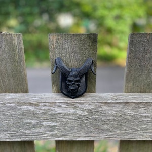 以下が含まれることがあります： 木製のフェンスに、角のある黒い3Dプリントの頭蓋骨が置かれています
