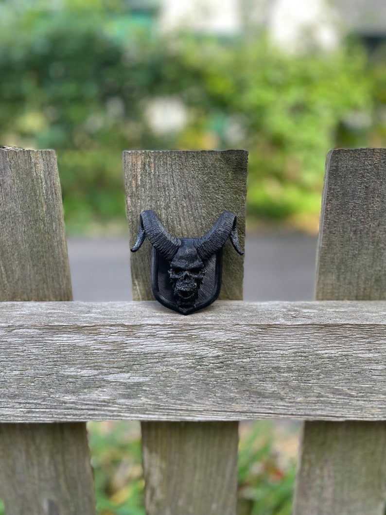 以下が含まれることがあります： 木製のフェンスに、角のある黒い3Dプリントの頭蓋骨が置かれています