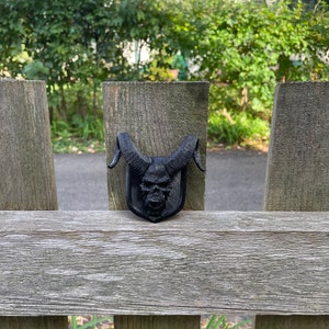以下が含まれることがあります： 木製のフェンスに取り付けられた、角のある黒い3Dプリントの頭蓋骨