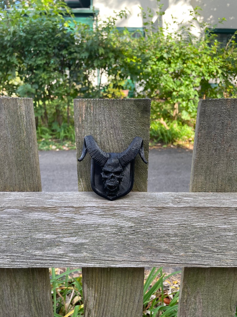 以下が含まれることがあります： 木製のフェンスに取り付けられた、角のある黒い3Dプリントの頭蓋骨