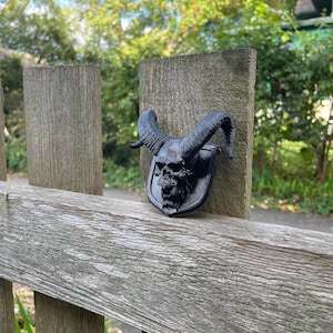 以下が含まれることがあります： 木製のフェンスの支柱に取り付けられた、角のある黒い3Dプリントの頭蓋骨