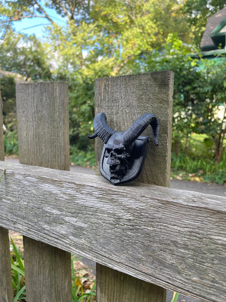 以下が含まれることがあります： 木製のフェンスの支柱に取り付けられた、角のある黒い3Dプリントの頭蓋骨