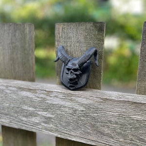 以下が含まれることがあります： 木製のフェンスに取り付けられた、角のある黒い頭蓋骨。頭蓋骨は、質感のある表面で詳細に作られており、プラスチック製のように見える。