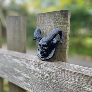 以下が含まれることがあります： 木製のフェンスの支柱に取り付けられた、角のある黒い3Dプリントの頭蓋骨