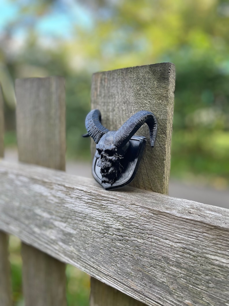 以下が含まれることがあります： 木製のフェンスの支柱に取り付けられた、角のある黒い3Dプリントの頭蓋骨