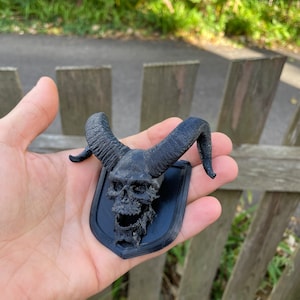 以下が含まれることがあります： 黒い3Dプリントされた頭蓋骨に角が取り付けられ、黒いプレートにマウントされています。頭蓋骨は詳細なデザインで、口が開いています。