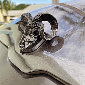 Pode incluir: Um ornamento de capô de carro com um crânio de carneiro preto. O crânio tem chifres detalhados e é montado em uma superfície escura. Um acessório decorativo para carros.
