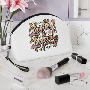 Könnte beinhalten: Eine weiße Kosmetiktasche mit schwarzem Reißverschluss und Handschlaufe. Die Tasche trägt den Text "Lashes & Leopard" in einem rosa-goldenen Design. Make-up-Pinsel und Lippenstifte sind auf einer Marmoroberfläche angeordnet.