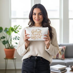 Könnte beinhalten: Eine weiße Kosmetiktasche mit goldenem Text, der lautet: "Girls just want to lash out!" Die Tasche hat auch eine schwarze Wimpernbürsten-Grafik. Die Tasche wird von einer Person gehalten, die einen cremefarbenen Pullover und eine schwarze Jeans trägt.
