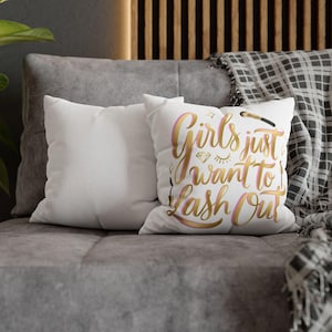 Op de afbeelding: Twee witte vierkante kussens op een grijze bank. Eén kussen heeft de tekst "Girls just want to lash out" in gouden en roze letters, met een diamant, wimpers en een make-up kwast.