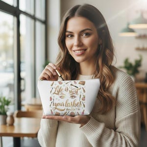 Könnte beinhalten: Weiße Kosmetiktasche mit goldenem Reißverschluss, verziert mit Make-up-Artikeln und dem Text "Life is short, your lashes shouldn't be." Die Tasche ist mit Make-up-Tools gefüllt.