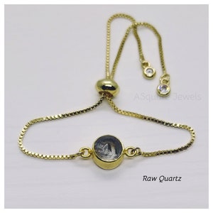 May include: Gold-toned adjustable bracelet featuring a raw quartz stone. The bracelet has a delicate chain and a sliding bead for adjustment. The quartz is set in a gold bezel. The text "Raw Quartz" is visible.