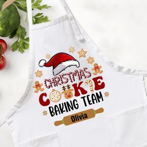 May include: White apron with a Christmas-themed design. The apron features a Santa hat, gingerbread men, and the text "Christmas Cookie Baking Team." A rolling pin graphic with the name "Olivia" is also included.