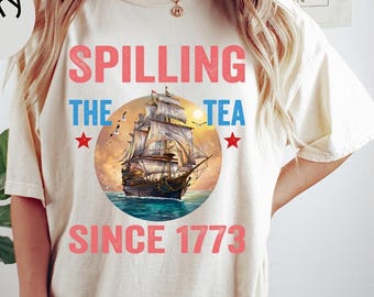 May include: Ivory-colored t-shirt with the text "Spilling the Tea Since 1773" in red and blue. The design features a detailed illustration of a sailing ship inside a circle. The shirt is a casual, comfortable style.