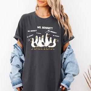 A woman wearing a gray t-shirt with a graphic of seagulls and text that says "MS. BENNETT" above them.